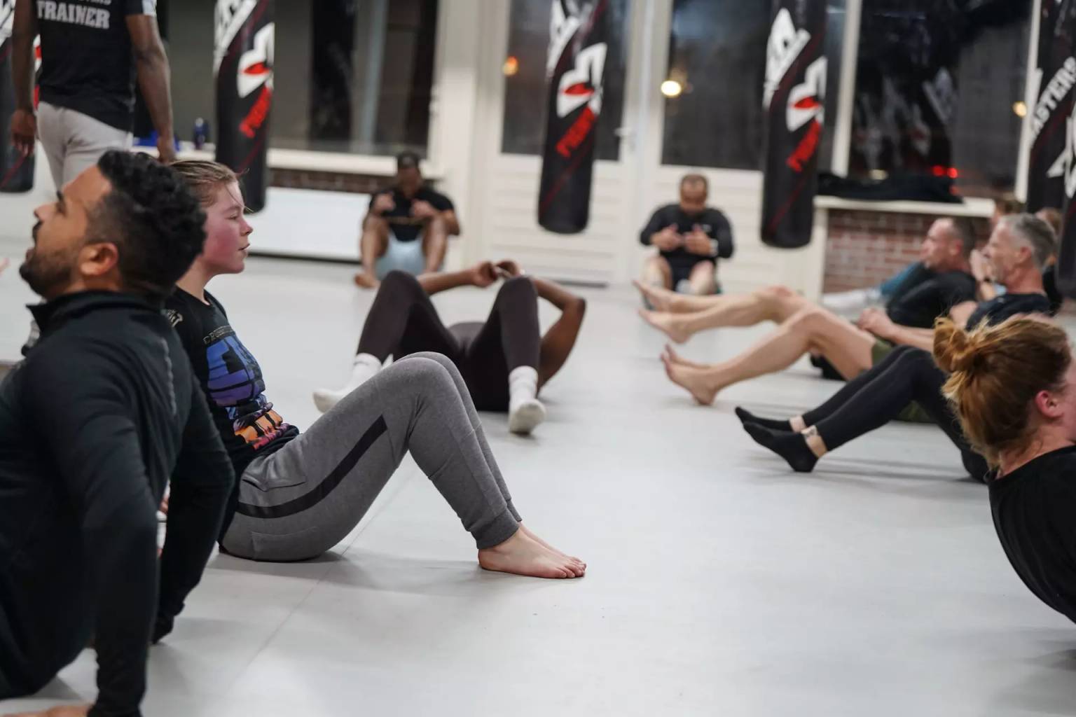 Een groep mensen die deelnemen aan een fitnessles gericht op vechtsport en sit-ups uitvoeren op de sportschoolvloer van Fightmasters omringd door bokszakken.