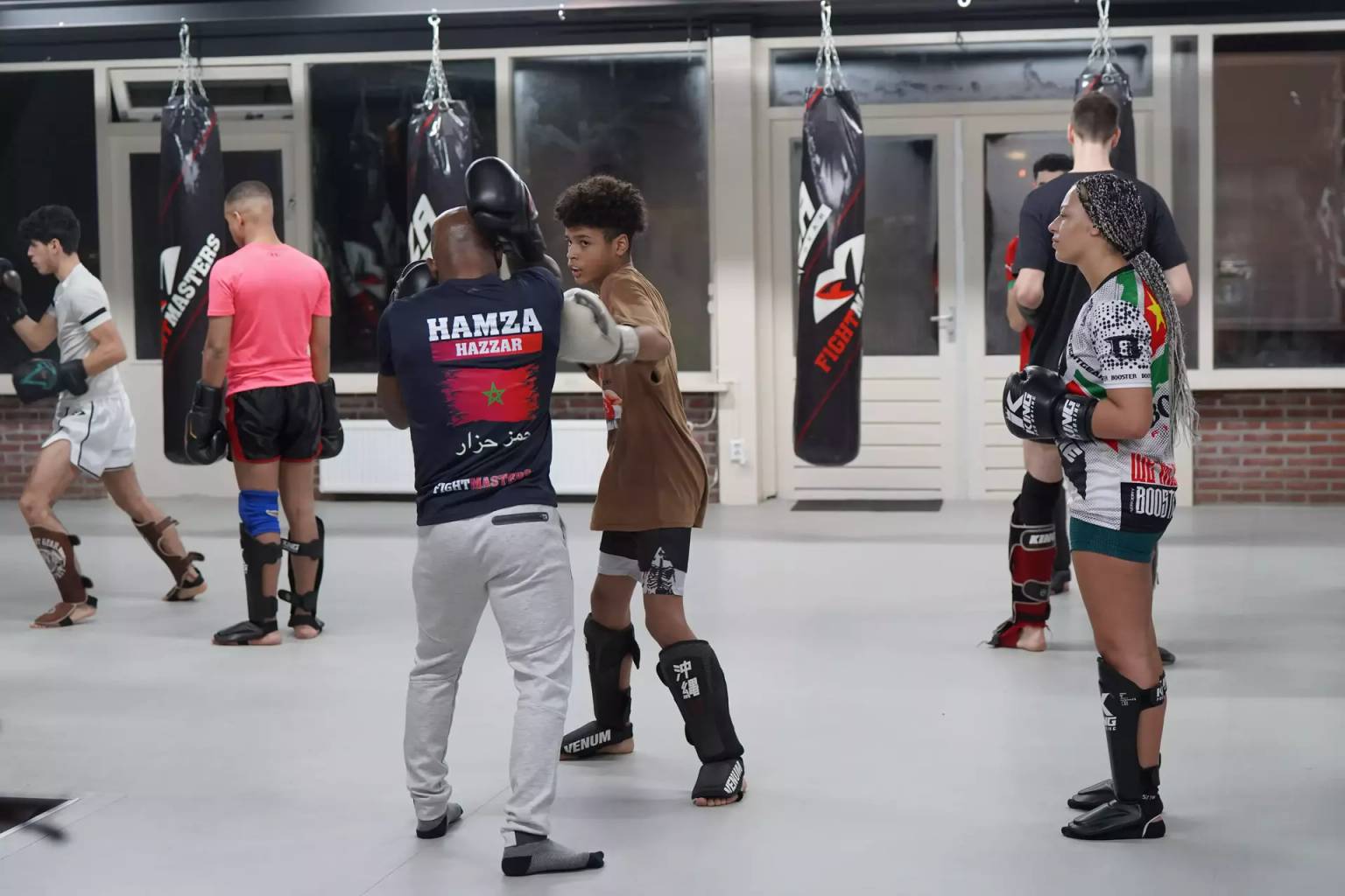 Een groep mensen die beschermende kleding dragen, neemt deel aan een kickbokstraining in sportschool Fightmasters, waarbij twee individuen op de voorgrond stoten oefenen en anderen op de achtergrond sparren.