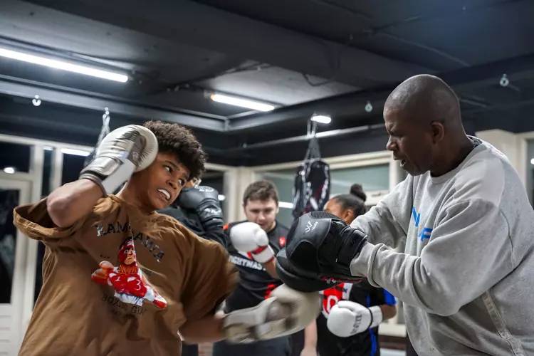 Een jonge man met bokshandschoenen oefent zijn stoten met een trainer in sportschool Fightmasters. Op de achtergrond zijn andere personen en boksuitrusting zichtbaar.