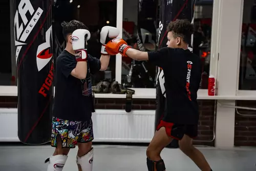 Twee mensen beoefenen boksen in sportschool Fightmasters. De een draagt een felgekleurde korte broek en blokkeert, terwijl de ander rode handschoenen draagt en een stoot uitdeelt.