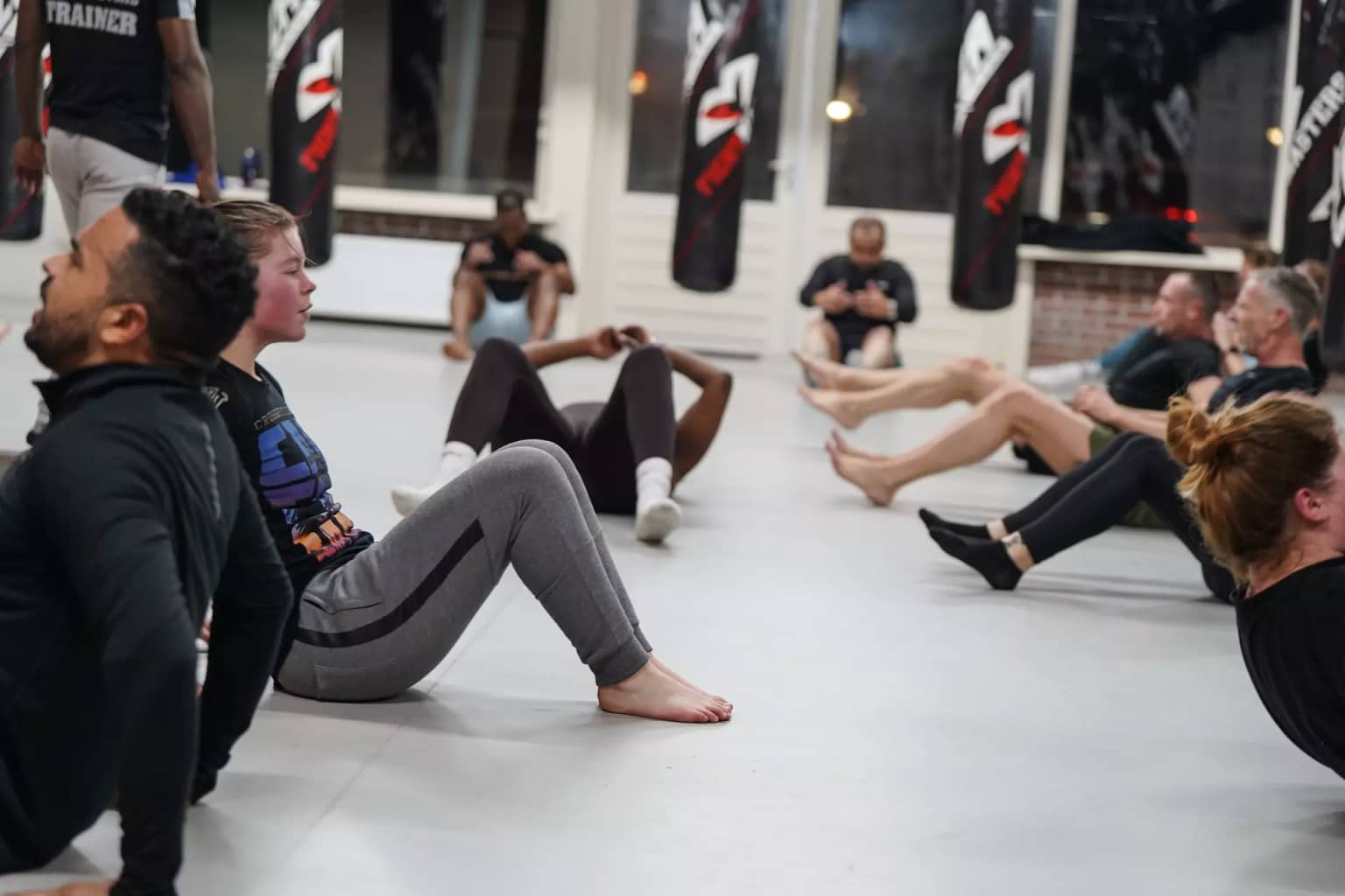 Een groep mensen in trainingskleding ligt op de grond in een sportschool en doet sit-up-oefeningen. Op de achtergrond zijn bokszakken en ramen zichtbaar.