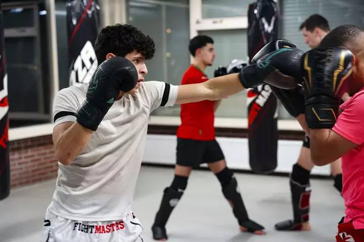 Een jonge man met bokshandschoenen oefent boksen door een stoot te geven aan een trainer die pads vasthoudt in op de sportschool van Fightmasters. Twee andere personen op de achtergrond zijn bezig met techniekoefeningen.