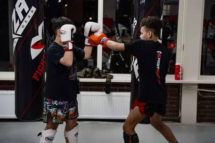 Twee jeugdvechters oefenen techniek in sportschool Fightmasters, elk met beschermende handschoenen en uitrusting. De ene persoon deelt een klap uit, terwijl de ander deze blokkeert.