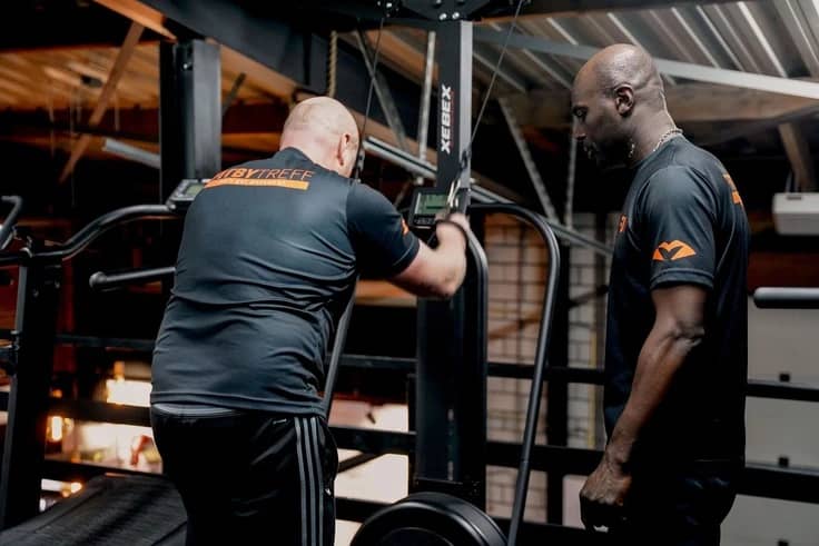 Twee mannen in sportkleding gebruiken een verticale klimmachine in sportschool Fightmasters. De ene man voert de oefening uit, terwijl de personal trainer toekijkt.